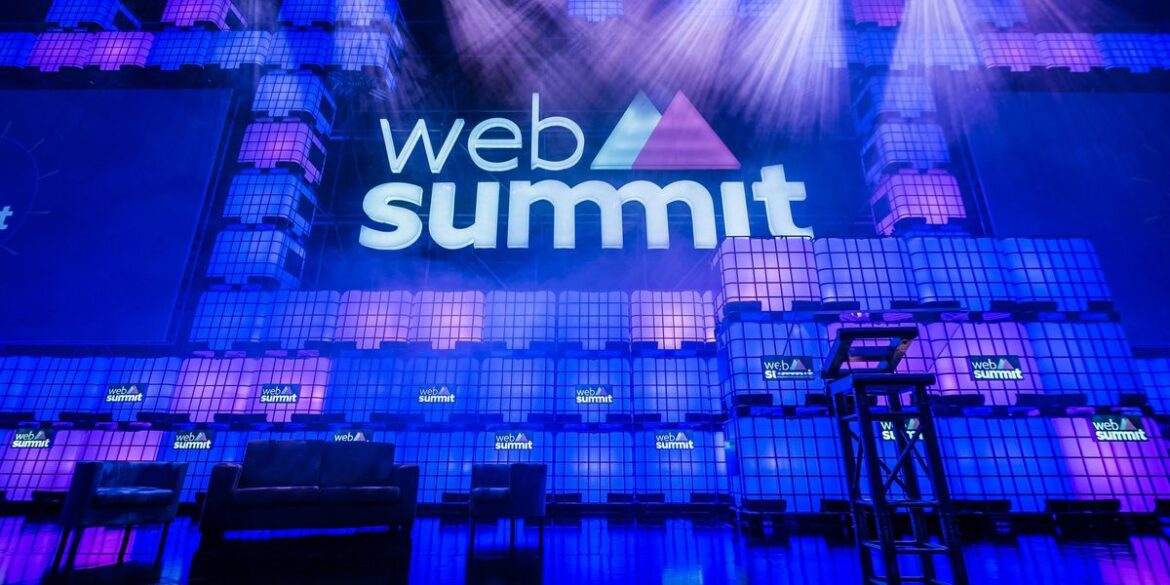 Stage with a purple and blue lit backdrop featuring web summit text with empty chairs and podium bel.jpeg