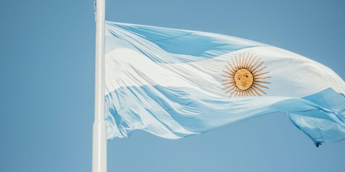 Argentinian flag waving on a pole against a clear blue sky displaying a sun emblem in the center.jpg