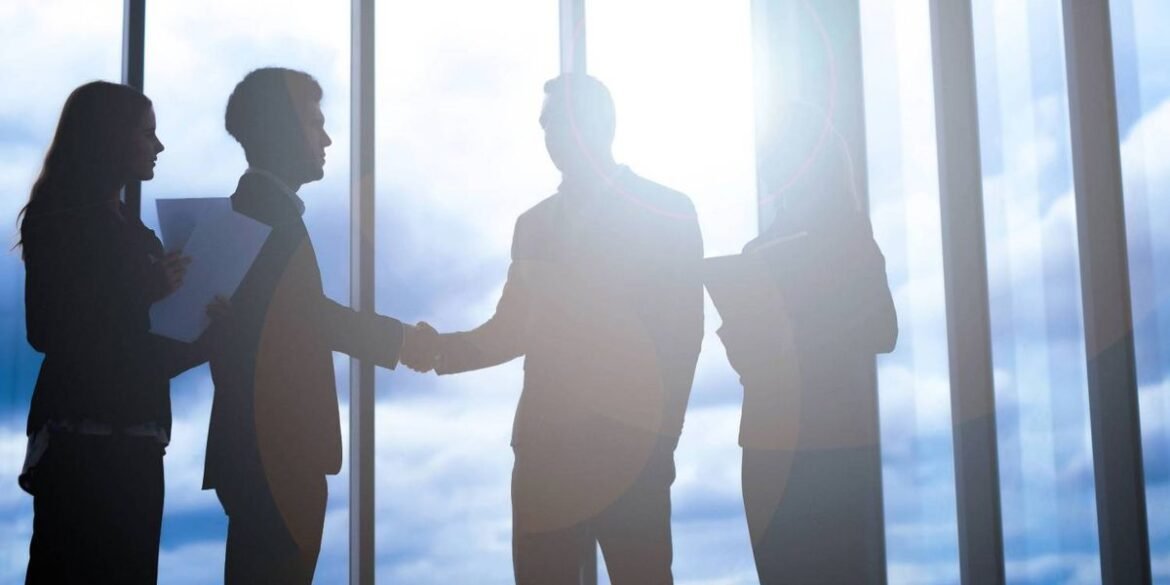 Silhouettes of four people two shaking hands stand in front of large windows with a bright sky backg.jpeg